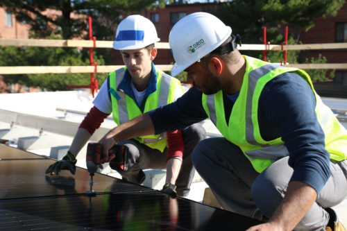 Solar installers wearing white helmets and reflective vests use a drill to fix the panels to the roof.